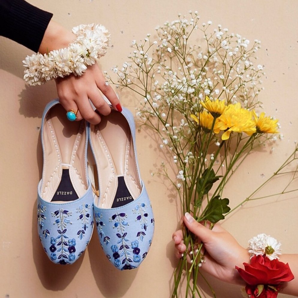 Light blue embroidered khussa shoes for women held by hand, with floral jewelry and fresh flowers