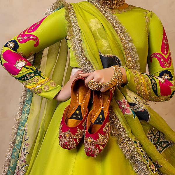 Woman in vibrant embroidered dress holding red gold embellished khussa shoes, traditional Pakistani footwear.