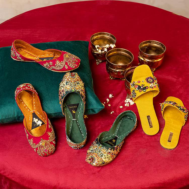 Embroidered women's khussa and kolhapuri chappal shoes in red, green, and yellow on a red velvet background with decorative brass pots.